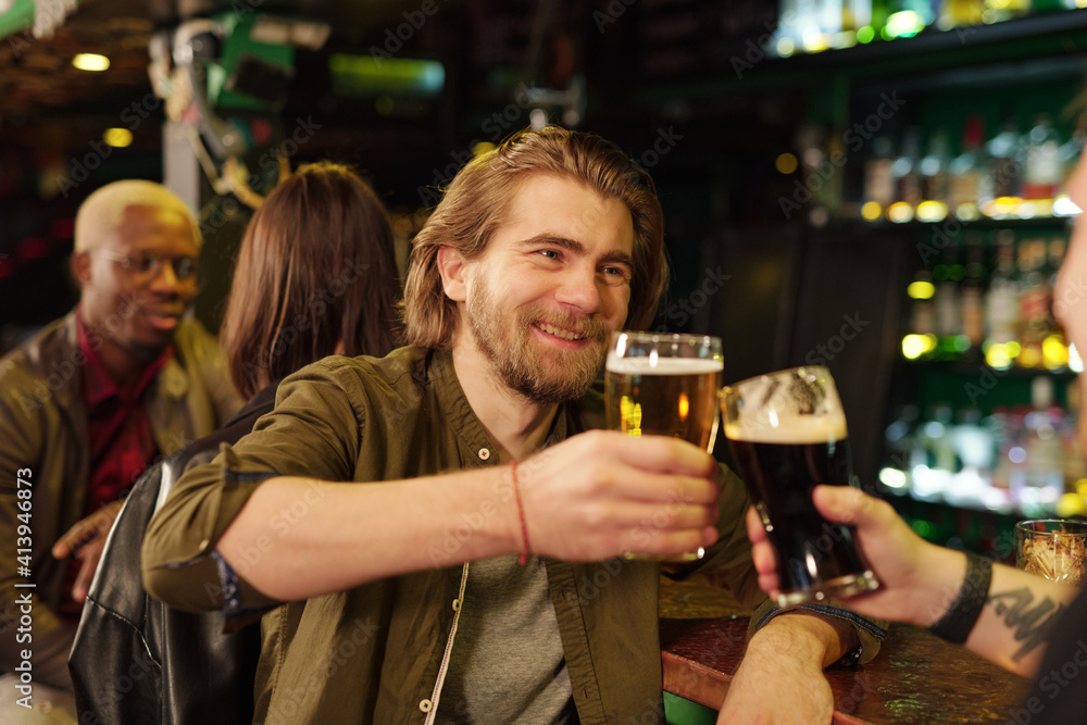 Happy young bearded man looking at friend while clinking with glasses of beer