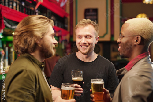 Three young cheerful intercultural men looking at one another while having beer