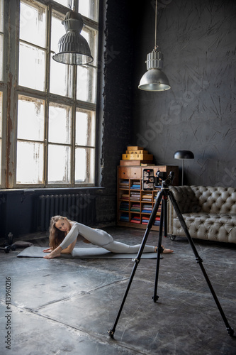 Young beautiful woman doing stretching exercises in loft style room. She conducts training by recording the broadcast on her smartphone. Sports during a pandemic, online gymnastics and yoga classes