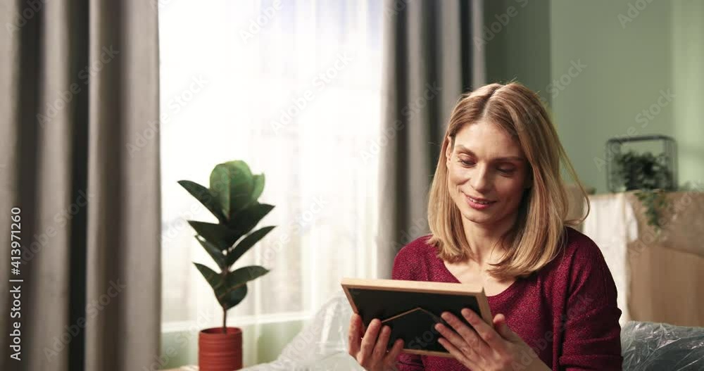 Beautiful happy woman sitting on sofa among cardboard boxes in new apartment and looking at photo in frame. Beautiful happy woman sitting on sofa among cardboard boxes in new apartment and looking at photo in frame.