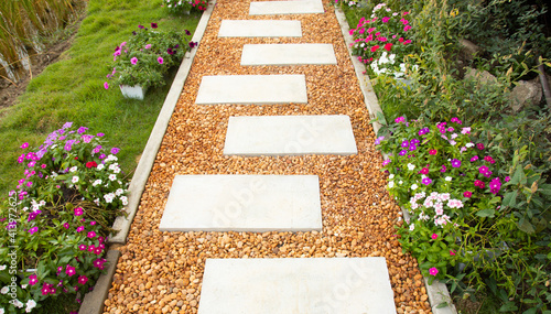 Selective focus shot of white stone path with yellow and brown gravel in colorful flower garden and green grass shows beautiful landscape of summer season. It is a gorgeous background of walkway