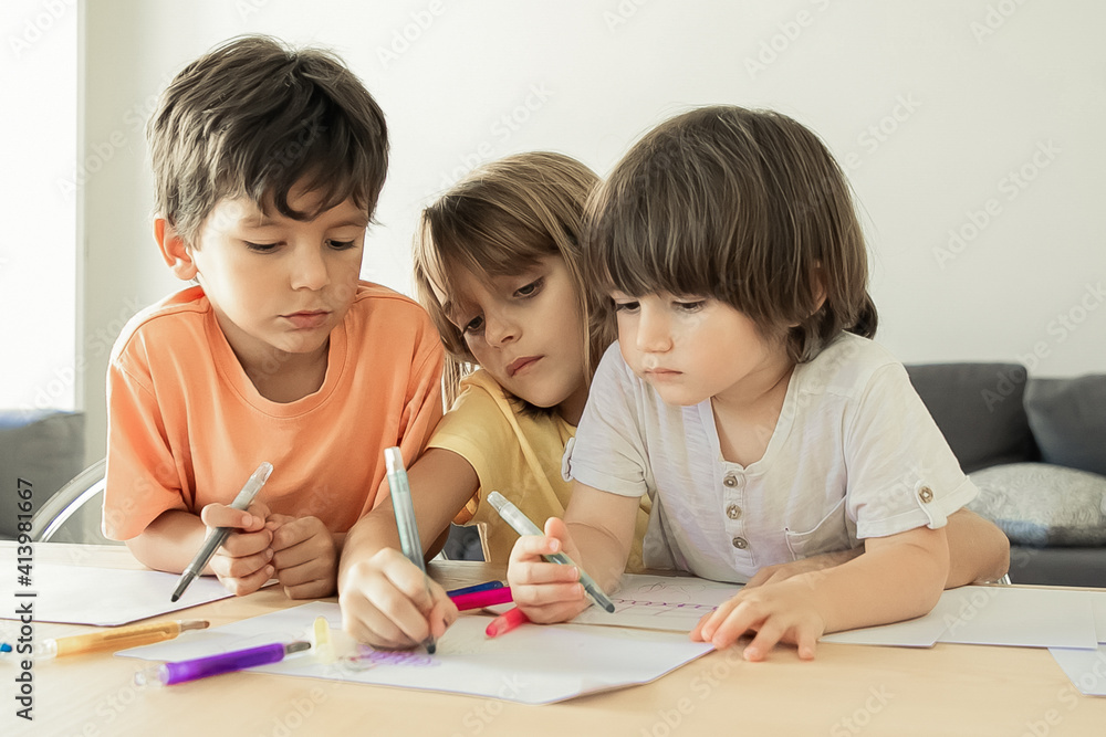 Thoughtful children painting with markers in living room. Caucasian lovely boys and blonde girl sitting at table, drawing on paper and playing together. Childhood, creativity and weekend concept Thoughtful children painting with markers in living room. Caucasian lovely boys and blonde girl sitting at table, drawing on paper and playing together. Childhood, creativity and weekend concept