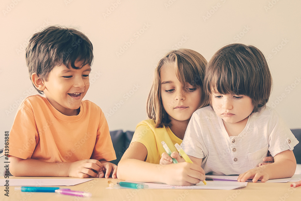 Happy children painting with markers in living room. Lovely little boys and blonde girl sitting at table, drawing on paper with pens and playing at home. Childhood, creativity and weekend concept