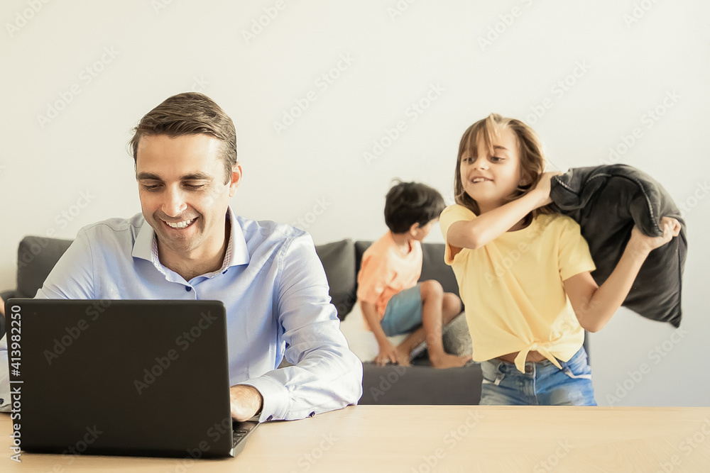 Cheerful dad chatting via laptop and kids playing with pillows near him. Caucasian father working at home during school vacations. Happy children on background. Family and digital technology concept Cheerful dad chatting via laptop and kids playing with pillows near him. Caucasian father working at home during school vacations. Happy children on background. Family and digital technology concept