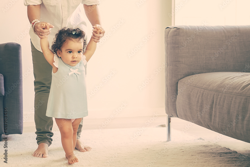 Focused baby girl in pale blue dress holding moms hands and trying to walk at home. Full length. Parenthood and childhood concept - obrazy, fototapety, plakaty 