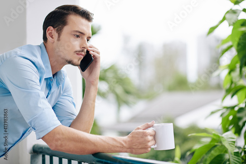 Frowning pensive young man drinking coffee on apartment balcony and talking on phone with friend or relative