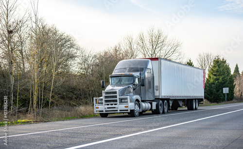 Gray stylish big rig semi truck with aluminum grille guard and dry van semi trailer standing on the road shoulder out of service