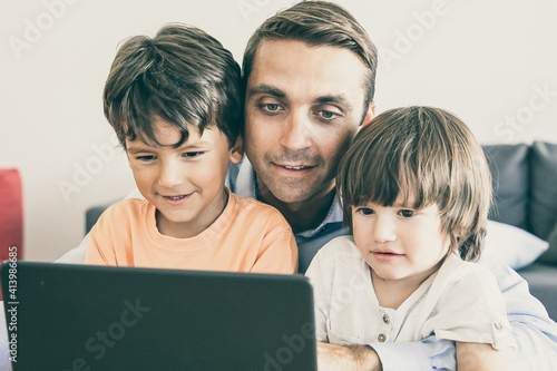 Happy middle-aged dad hugging sons and watching movie on laptop. Cute little boys sitting at table with father, looking at screen and smiling. Fatherhood, childhood and digital technology concept