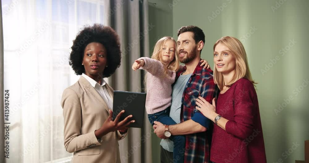 Cheerful Caucasian couple with little child standing in new empty house talking to African American female real estate sales manager who is using tablet showing purchase details. Buying home concept Cheerful Caucasian couple with little child standing in new empty house talking to African American female real estate sales manager who is using tablet showing purchase details. Buying home concept