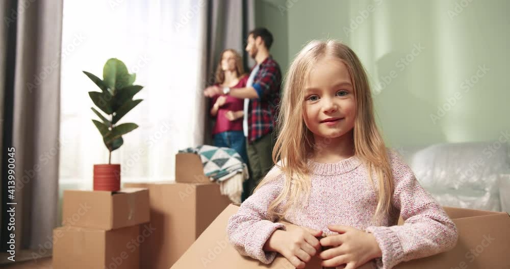 Close up of joyful Caucasian little girl kid sitting in big carton box having fun playing and smiling to camera while parents standing and looking in window behind. Unpacking on relocation day Close up of joyful Caucasian little girl kid sitting in big carton box having fun playing and smiling to camera while parents standing and looking in window behind. Unpacking on relocation day