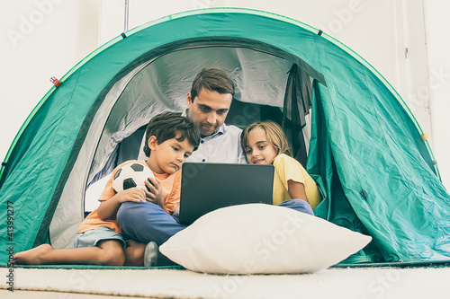 Caring dad sitting cross-legged with kids in tent at home and looking at laptop screen. Adorable children watching movie on computer with father. Childhood, family time and weekend concept