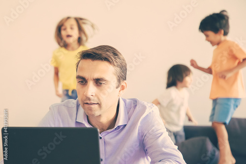 Serious Caucasian dad working via laptop and kids jumping on background. Concentrated father using computer and kids playing on sofa. Selective focus. Childhood and digital technology concept © Mangostar