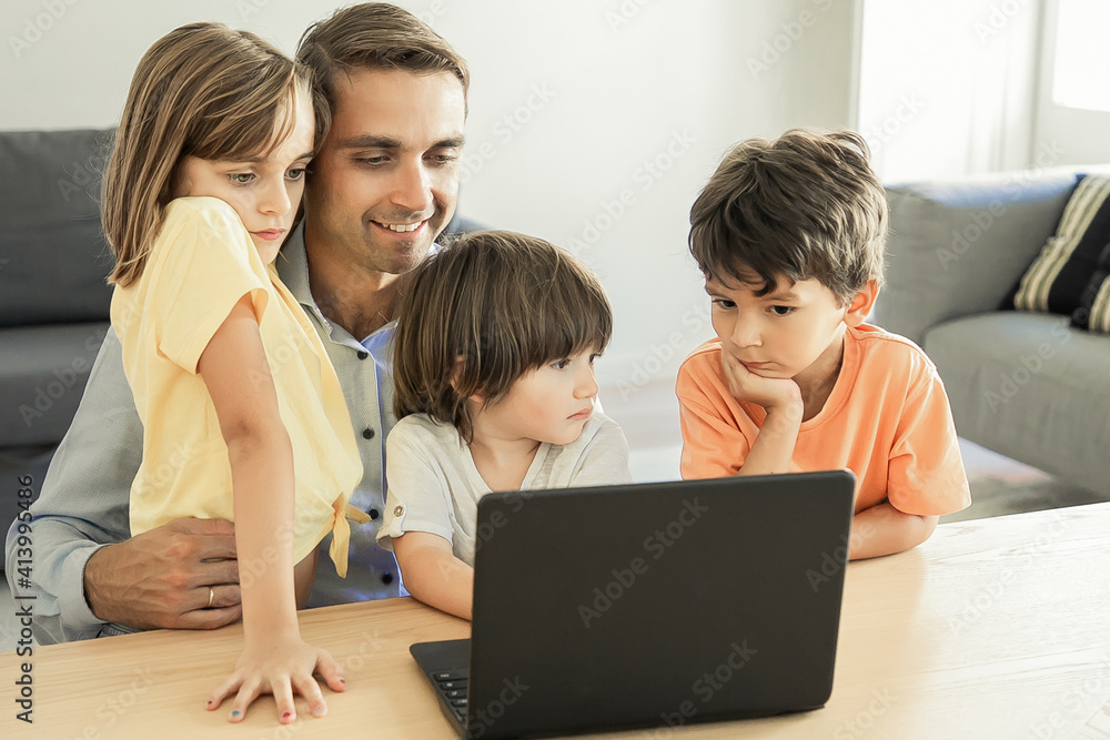 Happy dad sitting at table, embracing kids and using laptop. Caucasian middle-aged father browsing in internet with lovely children. Childhood, fatherhood and digital technology concept