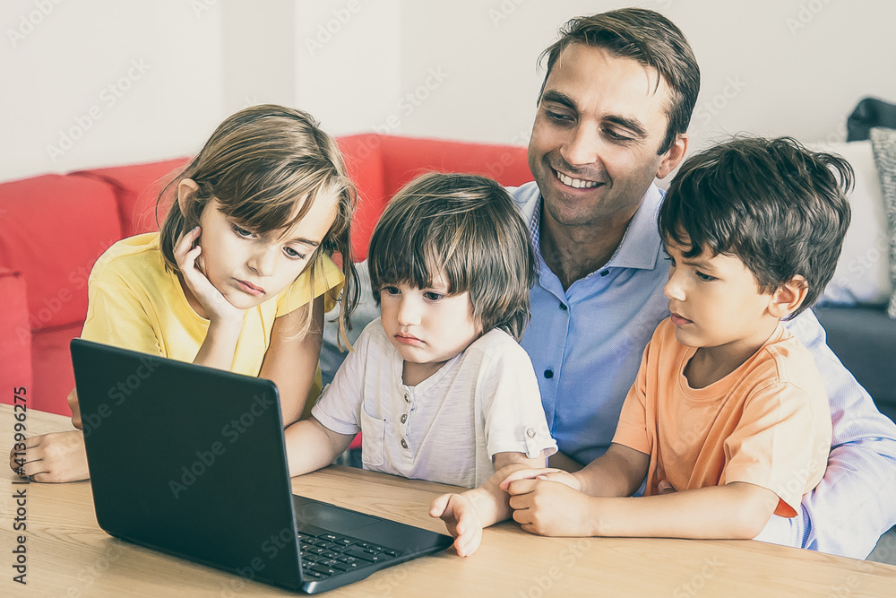 Cheerful dad and pensive kids watching movie via laptop together during weekend. Happy father sitting at table with children in living room. Fatherhood, childhood and digital technology concept Cheerful dad and pensive kids watching movie via laptop together during weekend. Happy father sitting at table with children in living room. Fatherhood, childhood and digital technology concept
