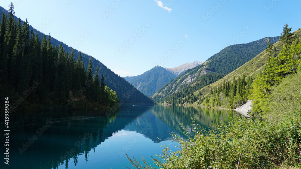 Kolsay lake among green hills and mountains. The mountain lake is surrounded by green forest, tall coniferous trees, grass and bushes. Clean water is like a mirror. Tourists swim on boats. Kazakhstan