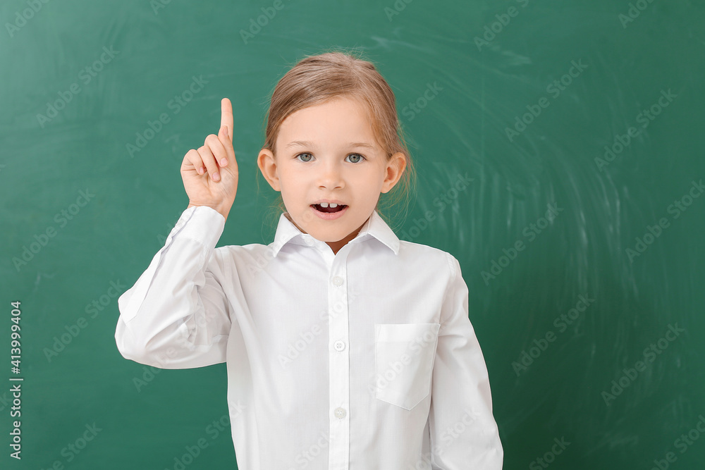 Little schoolgirl with raised index finger near chalkboard in classroom