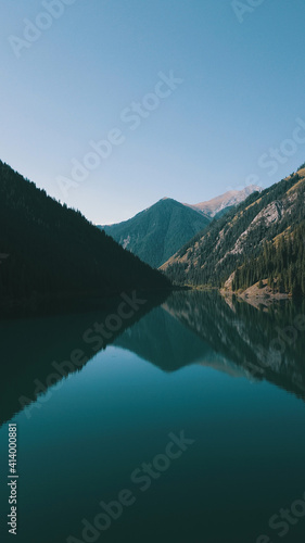 Kolsay lake among green hills and mountains. The mountain lake is surrounded by green forest  tall coniferous trees  grass and bushes. Clean water is like a mirror. Tourists swim on boats. Kazakhstan