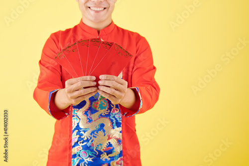 Cropped image of cheerful young smiling man holding red lucky money envelopes, isolated on yellow background