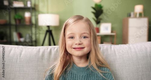 Close up portrait of Caucasian little adorable happy child girl sitting on sofa in room, looking at camera and smiling in positive mood. Happy childhood. Small cute pre-school kid at home alone photo