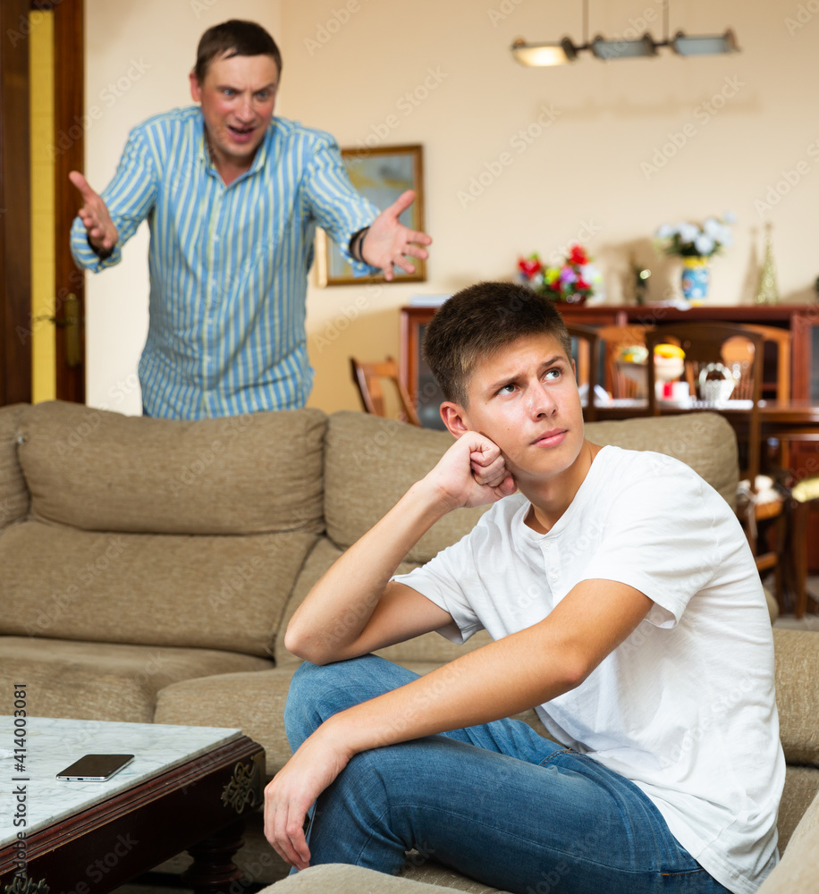 Frowning teen boy sitting at home while father berating him ..