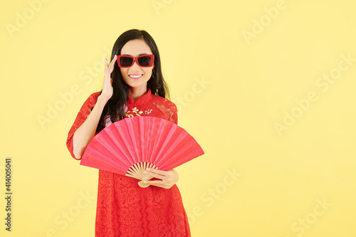 Cheerful beautiful young Asian woman in lace red dress putting on sunglasses and holding paper fan