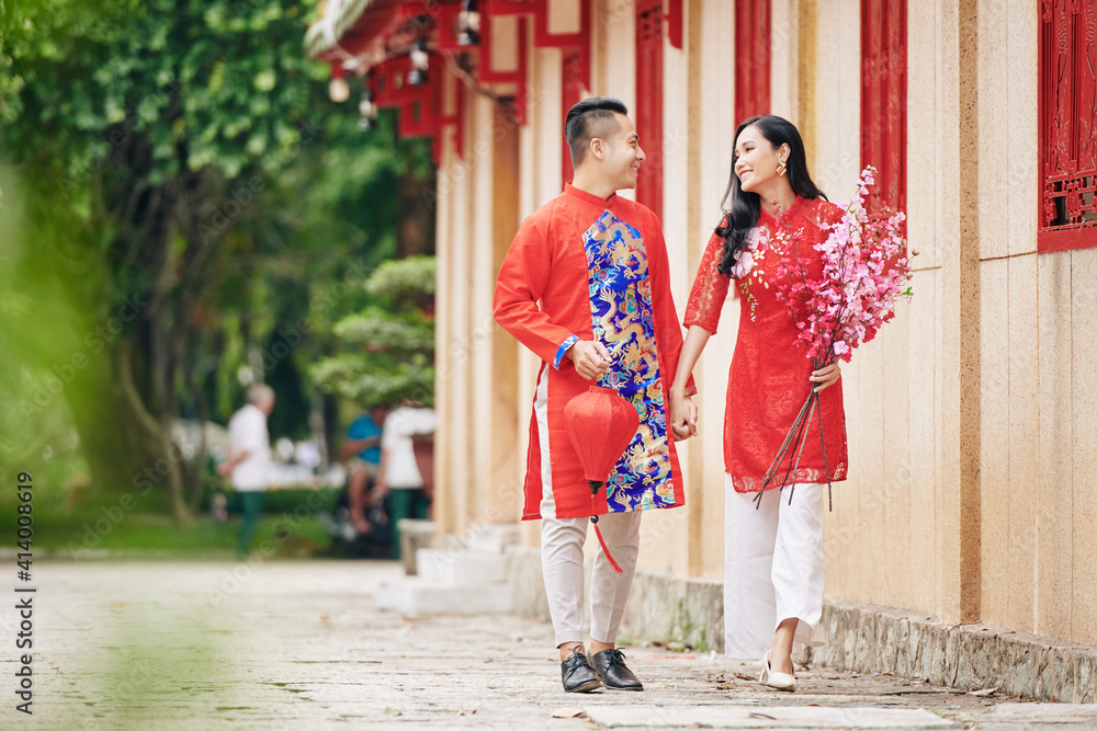 Happy young Vietnamese boyfriend and girlfriend with silk lantern and blooming peach branches holding hands when walking on street