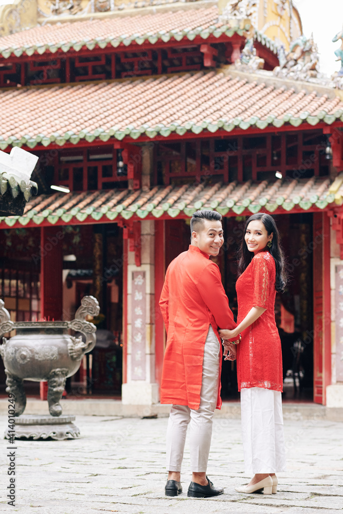 Happy young Asian couple in traditional dresses walking to Buddhism temple, turning back and smiling at camera