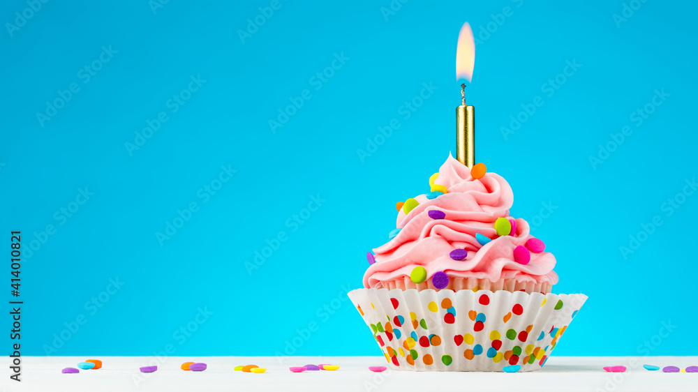 Birthday cupcake. Tasty cupcakes with pink cream icing and colored sprinkles. Burning candle in a cupcake. Sweet delicious dessert on natural white wooden table and blue background. Happy Birthday. Birthday cupcake. Tasty cupcakes with pink cream icing and colored sprinkles. Burning candle in a cupcake. Sweet delicious dessert on natural white wooden table and blue background. Happy Birthday.