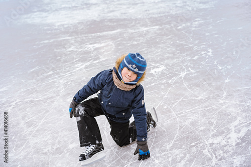 cute caucasian preschooler boy learn to skate on ice rink. Kid wearing winter jacket, hood hat and neckwarmer standing on one knee. Image with selective focus photo