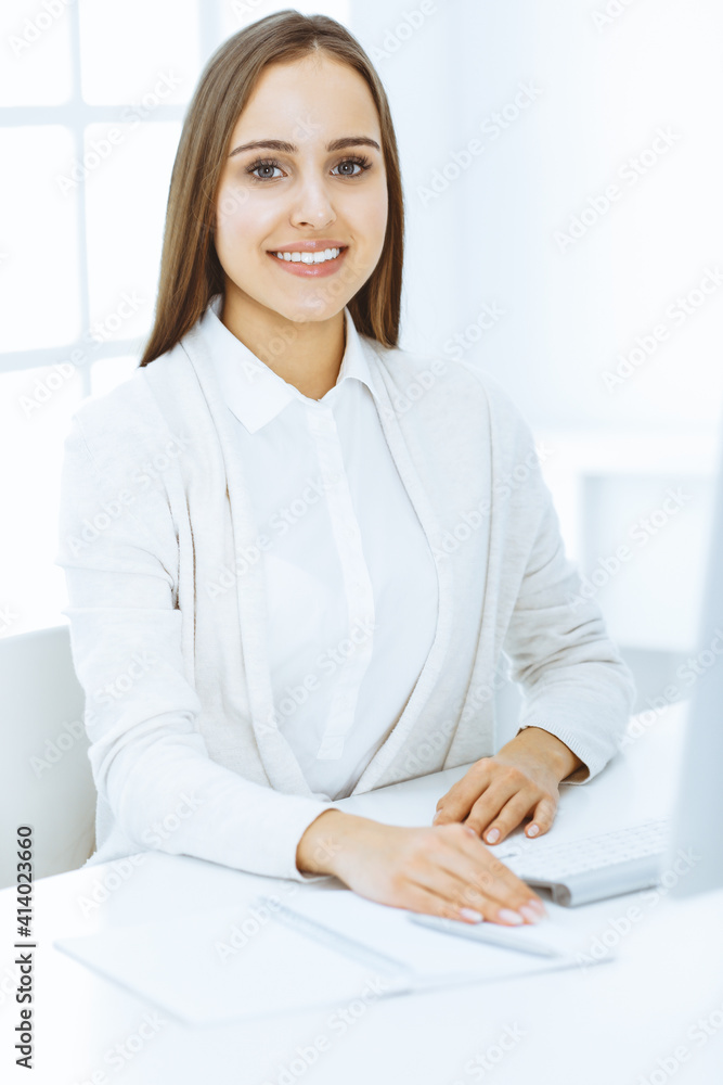 Young cheerful woman sitting at the desk with computer and looking at camera in white colored office. Looks like student girl or business lady. Headshot Young cheerful woman sitting at the desk with computer and looking at camera in white colored office. Looks like student girl or business lady. Headshot
