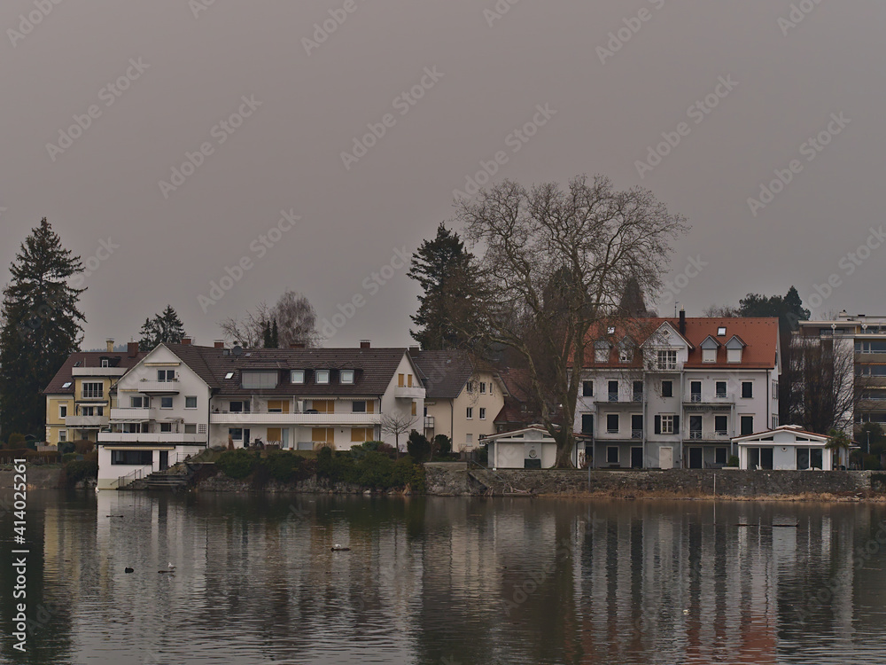 Residential buildings on the shore of Lake Constance in the center of Lindau, Germany with water reflections and bare trees in winter season on foggy day.