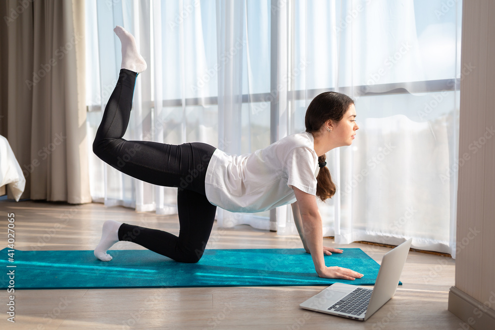 Young beautiful woman in sports clothes, doing yoga online lessons on a laptop. Indoor. Side view. The concept of online training at home Young beautiful woman in sports clothes, doing yoga online lessons on a laptop. Indoor. Side view. The concept of online training at home