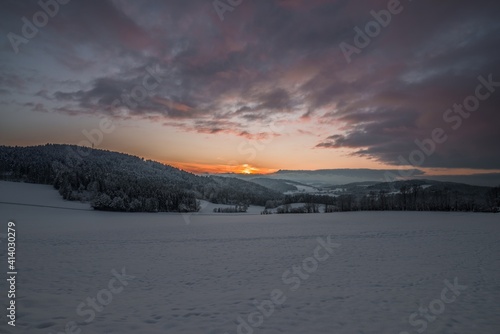 Landschaft während Sonnenuntergang im Winter mit Schnee und Eis und Berge im Hintergrund bei Grafenau im Bayerischer Wald mit Wolken und Sonnenstrahlen, Deutschland