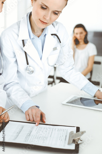 Two woman-doctors at work with patient at background. Female physicians filling up medical documents or prescription while standing in hospital reception desk. Data in medicine