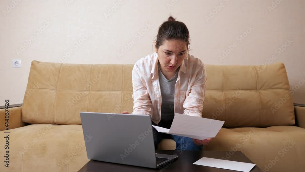Emotional indignant woman with bill paper calculates payments on modern laptop sitting on beige comfortable couch in light living room Emotional indignant woman with bill paper calculates payments on modern laptop sitting on beige comfortable couch in light living room