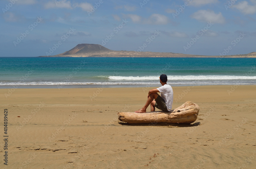 Unrecognizable traveller sitting on a wooden log on the beach watching the sea