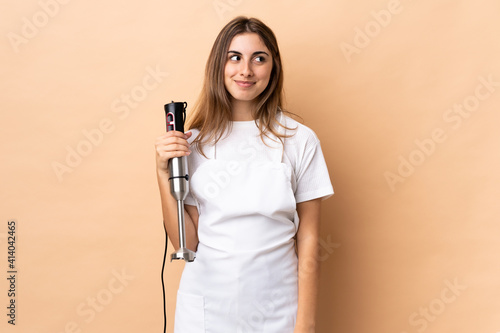 Woman using hand blender over isolated background making doubts gesture looking side