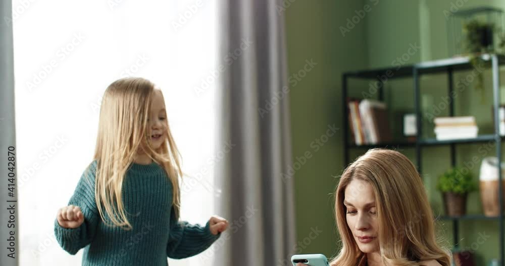 Focused mom working from home answering email from smartphone while daughter jumps on couch next to her. Focused mom working from home answering email from smartphone while daughter jumps on couch next to her.