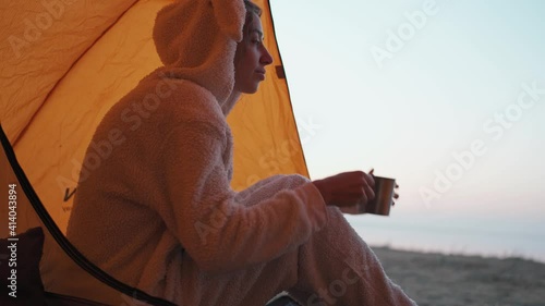 pretty woman in funny pajama like bunny sitting at orange tent entry, drinking hot coffee from metal mug and enjoying sunrise view at fresh cold morning in camping during travel photo