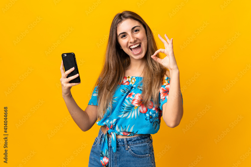 Young woman using mobile phone over isolated yellow background showing ok sign with fingers Young woman using mobile phone over isolated yellow background showing ok sign with fingers