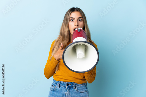 Young hispanic woman over isolated blue background shouting through a megaphone with surprised expression