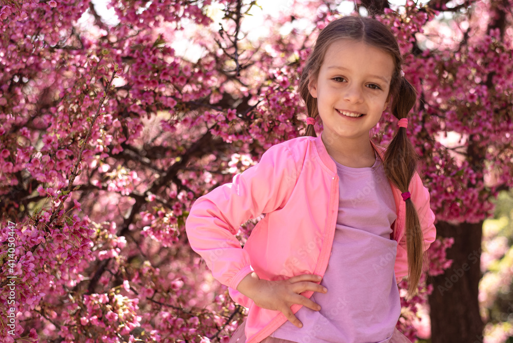 Little cute girl in garden on background of blooming pink cherry. Happy smile on her face. Pink jacket and lilac T-shirt go well with sakura flowers. Spring is time of blooming of nature. Outdoors. Little cute girl in garden on background of blooming pink cherry. Happy smile on her face. Pink jacket and lilac T-shirt go well with sakura flowers. Spring is time of blooming of nature. Outdoors.