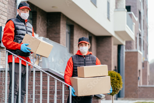 Two delivery man in masks and gloves workers in cap, red uniform walking and looking for address. Adult couriers delivering order in cardboard boxes. Delivery service, post and shipping concept