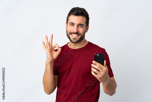 Young handsome man with beard using mobile phone isolated on white background showing ok sign with fingers
