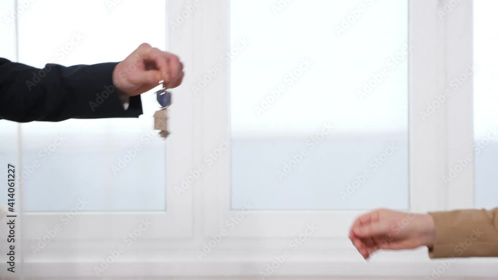 Businessman and young woman in office suits shake hands and man hands over keys to apartment near large window in light spacious room closeup Businessman and young woman in office suits shake hands and man hands over keys to apartment near large window in light spacious room closeup