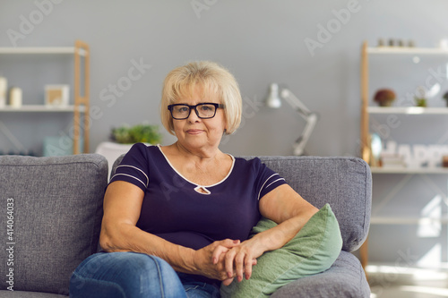 Positive elderly woman pensioner grandmother in jeans, t-shirt and glasses sitting on sofa at home and looking at camera with room interior background. Happy elderly people, retired women concept