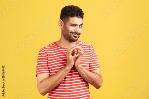Sneaky cunful man with beard in striped red t-shirt drumming fingers planning devious pranks, thinking over revenge plan, scheming. Indoor studio shot isolated on yellow background photo
