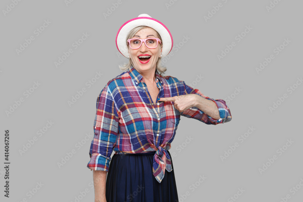 who? me? Portrait of surprised modern mature woman in casual style with hat and eyeglasses standing, looking with amazed face, pointing herself and asking. studio shot isolated on gray background. who? me? Portrait of surprised modern mature woman in casual style with hat and eyeglasses standing, looking with amazed face, pointing herself and asking. studio shot isolated on gray background.