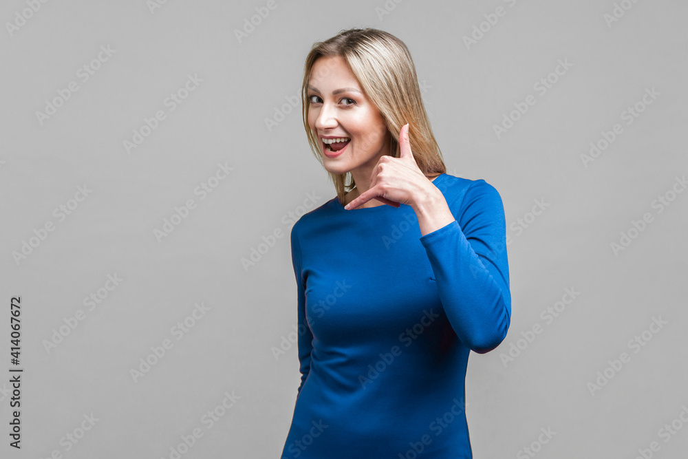 Call me back! Portrait of playful attractive woman in elegant tight blue dress doing talking on phone gesture, smiling and flirting at camera. indoor studio shot isolated on gray background Call me back! Portrait of playful attractive woman in elegant tight blue dress doing talking on phone gesture, smiling and flirting at camera. indoor studio shot isolated on gray background