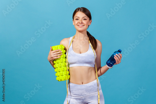 Slim self confident woman in white sportswear holding fitness roller and bottle with fresh table water, going on training at gym. Indoor studio shot isolated on blue background photo